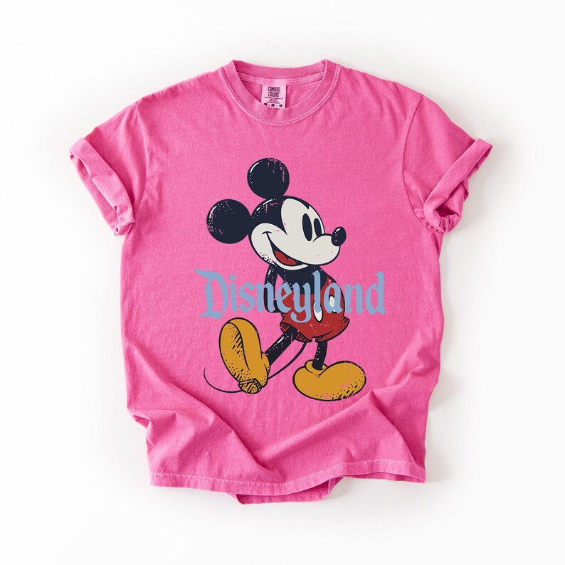 Puede incluir: Una camiseta rosa con un gr&aacute;fico de Mickey Mouse caminando y la palabra "Disneyland" en azul. Mickey Mouse es negro, blanco, rojo y amarillo. La camiseta tiene las mangas enrolladas.