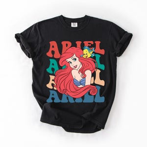 Op de afbeelding: Zwart T-shirt met een kleurrijke afbeelding van Ariel uit De Kleine Zeemeermin. Het ontwerp bevat Ariel's afbeelding, een gele vis en de naam "ARIEL" in retroletters. Een comfortabel, casual kledingstuk.