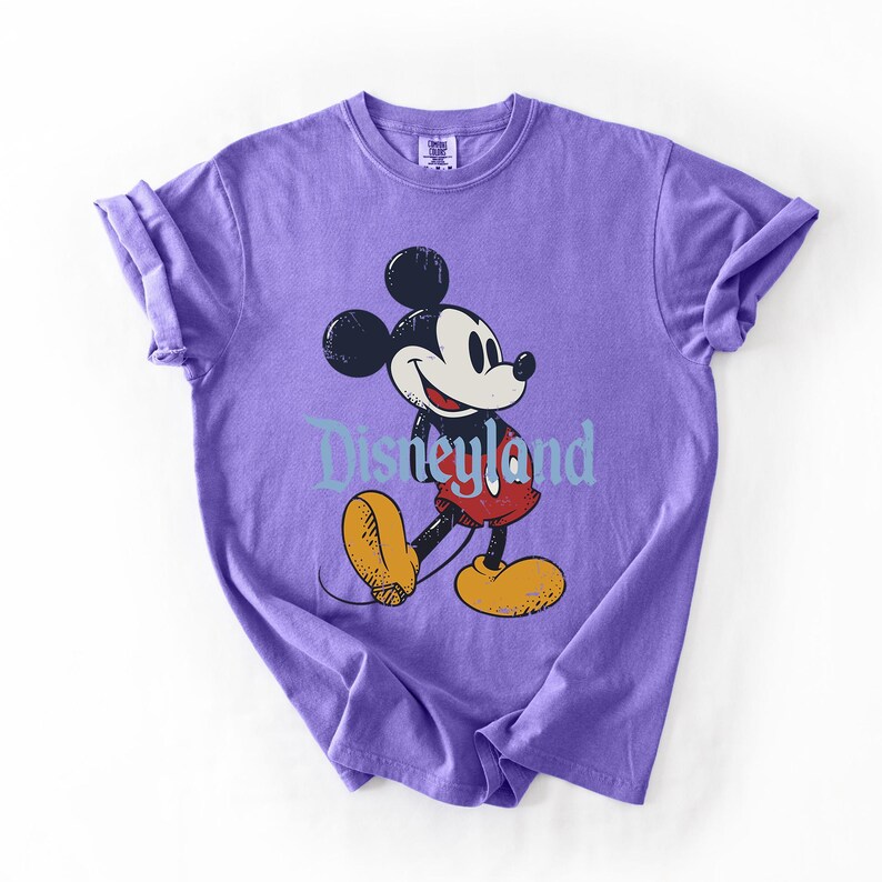 Puede incluir: Una camiseta morada con un gr&aacute;fico de Mickey Mouse y la palabra "Disneyland" en azul claro. Mickey se muestra en su pose cl&aacute;sica, con pantalones cortos rojos y zapatos amarillos. Las mangas de la camiseta est&aacute;n enrolladas.