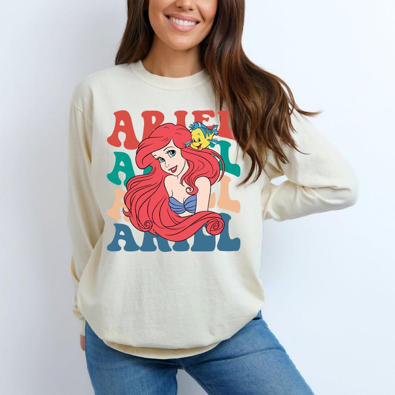 Op de afbeelding: Cr&egrave;mekleurige sweatshirt met lange mouwen met een afbeelding van Ariel uit De Kleine Zeemeermin. Het ontwerp bevat de naam "ARIEL" in kleurrijke, retro-stijl letters, met Ariel en Botje. De sweatshirt is gemaakt van een zacht materiaal.