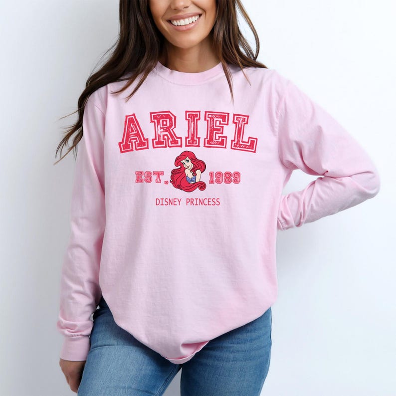 K&ouml;nnte beinhalten: Hellrosa Langarmshirt mit dem Namen "ARIEL" in roten Blockbuchstaben. Darunter eine Grafik von Arielle, der Disney-Prinzessin, mit dem Text "EST. 1989 DISNEY PRINCESS". Das Shirt ist aus weichem Material.