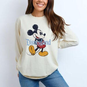 Puede incluir: Sudadera de manga larga color crema con un gr&aacute;fico desgastado de Mickey Mouse y la palabra "Disneyland" en escritura azul. El personaje de dibujos animados est&aacute; representado con su atuendo cl&aacute;sico con pantalones cortos rojos y zapatos amarillos.