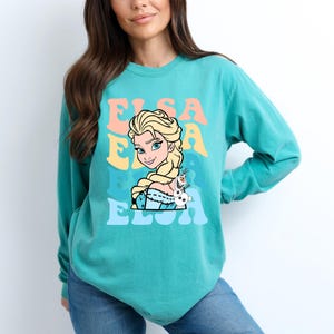 Puede incluir: Camiseta de manga larga color turquesa con un gr&aacute;fico de Elsa de Frozen, con la palabra "ELSA" en una fuente retro. El gr&aacute;fico incluye el retrato de Elsa y Olaf el mu&ntilde;eco de nieve. La camiseta es de material suave.