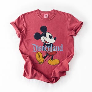 Puede incluir: Camiseta roja con un dibujo de Mickey Mouse y la palabra "Disneyland" en azul. Mickey Mouse est&aacute; representado en negro, blanco, amarillo y rojo. La camiseta tiene mangas enrolladas y cuello redondo.