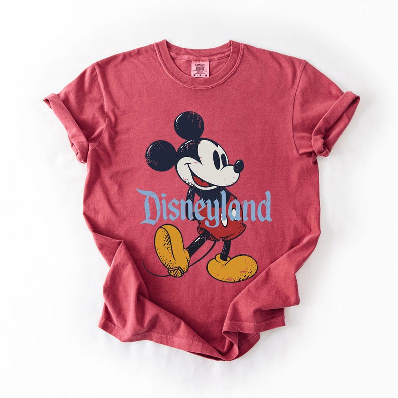 Puede incluir: Camiseta roja con un dibujo de Mickey Mouse y la palabra "Disneyland" en azul. Mickey Mouse est&aacute; representado en negro, blanco, amarillo y rojo. La camiseta tiene mangas enrolladas y cuello redondo.