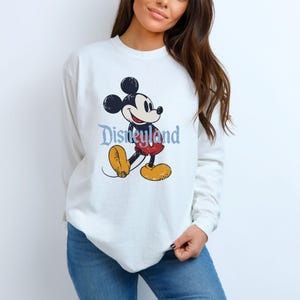 Puede incluir: Sudadera blanca de manga larga con un gr&aacute;fico desgastado de Mickey Mouse y la palabra "Disneyland" en azul. El dise&ntilde;o presenta a Mickey Mouse en su pose cl&aacute;sica. Los colores incluyen negro, rojo, amarillo y azul.