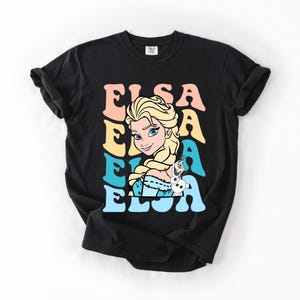 Puede incluir: Camiseta negra con un dise&ntilde;o retro de Elsa de Frozen. La palabra "ELSA" est&aacute; apilada en rosa, amarillo y azul. La imagen incluye a Elsa y Olaf el mu&ntilde;eco de nieve. El dise&ntilde;o es en tonos rosa, amarillo, azul y blanco.