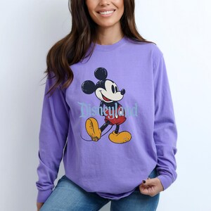Puede incluir: Sudadera de manga larga de color lavanda con un dise&ntilde;o de Mickey Mouse caminando. El texto "Disneyland" est&aacute; impreso debajo. El dise&ntilde;o incluye las orejas negras de Mickey, pantalones cortos rojos y zapatos amarillos.