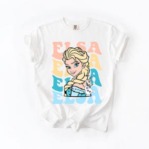 Puede incluir: Camiseta blanca con un gr&aacute;fico colorido de Elsa de Frozen, con el nombre "Elsa" en letras retro. El dise&ntilde;o incluye el retrato de Elsa y un peque&ntilde;o personaje de Olaf. Una camiseta divertida e informal.