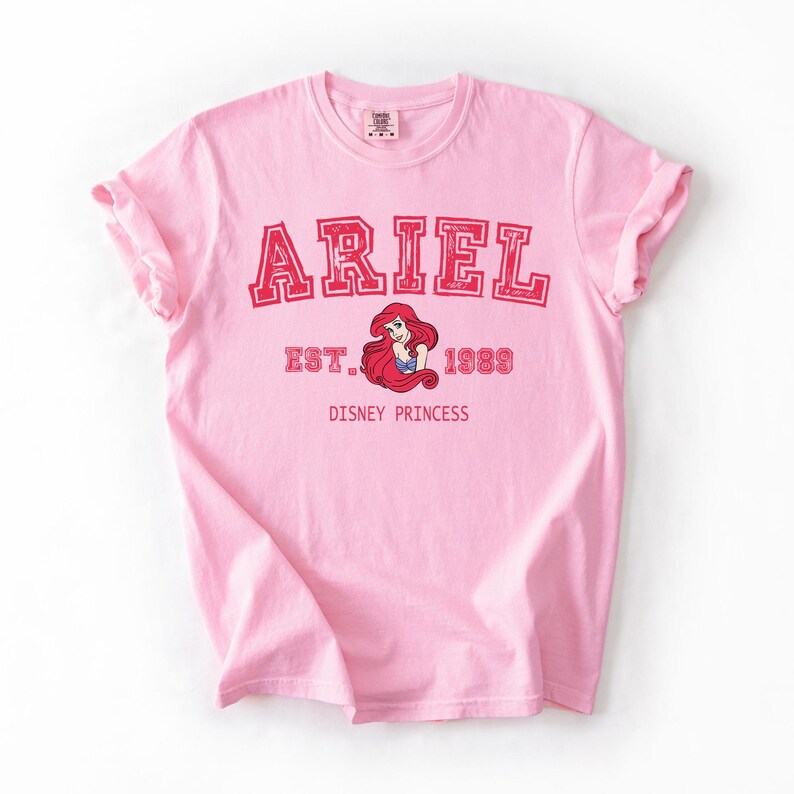 K&ouml;nnte beinhalten: Hellrosa T-Shirt mit dem Namen "ARIEL" in roten Blockbuchstaben. Darunter steht "EST. 1989" und eine Grafik von Arielle aus Disneys Arielle, die Meerjungfrau. Die Worte "DISNEY PRINCESS" sind unter der Grafik aufgedruckt.