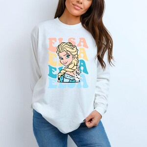 Puede incluir: Sudadera blanca de manga larga con un gr&aacute;fico colorido de Elsa de Frozen, con el nombre "Elsa" en colores pastel. El dise&ntilde;o incluye el retrato de Elsa y Olaf.