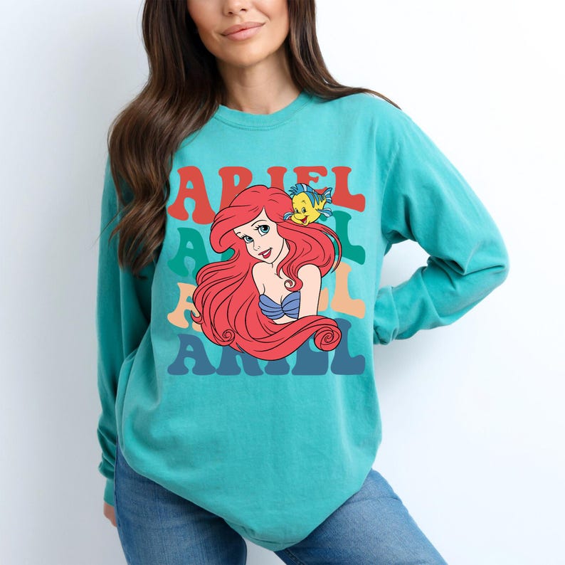 Op de afbeelding: Tealkleurig shirt met lange mouwen met een afbeelding van Ariel uit De Kleine Zeemeermin, met de naam "ARIEL" in kleurrijke retroletters. Het ontwerp bevat Ariels rode haar, een blauwe top en een gele vis.