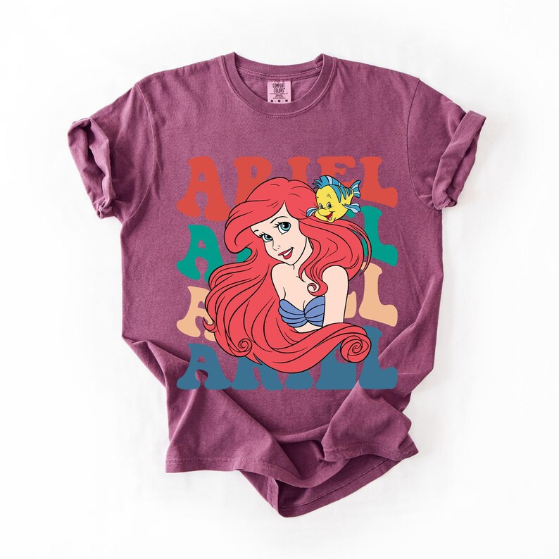 Op de afbeelding: Een bordeauxrood T-shirt met een kleurrijke afbeelding van Ariel uit De Kleine Zeemeermin. Het ontwerp bevat Ariel's afbeelding, een gele vis en de naam "ARIEL" in retro letters. Het shirt is gemaakt van zacht materiaal.