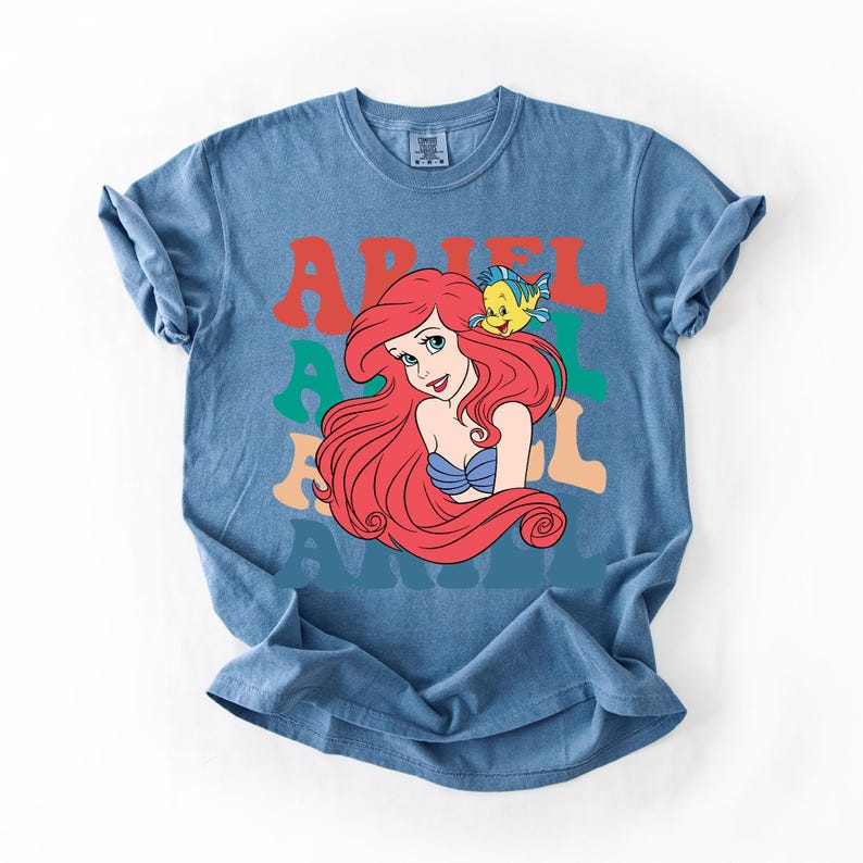 Op de afbeelding: Blauw T-shirt met een afbeelding van Ariel met rood haar en een gele vis. Het woord "ARIEL" is gedrukt in retro letters in rood, groen en oranje. Een leuk en kleurrijk ontwerp.