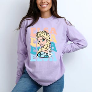Puede incluir: Una camiseta de manga larga de color lila claro con un gr&aacute;fico de Elsa de Frozen, y el nombre "ELSA" en una fuente retro. Elsa se muestra con cabello rubio y un vestido azul, con Olaf el mu&ntilde;eco de nieve a su lado.