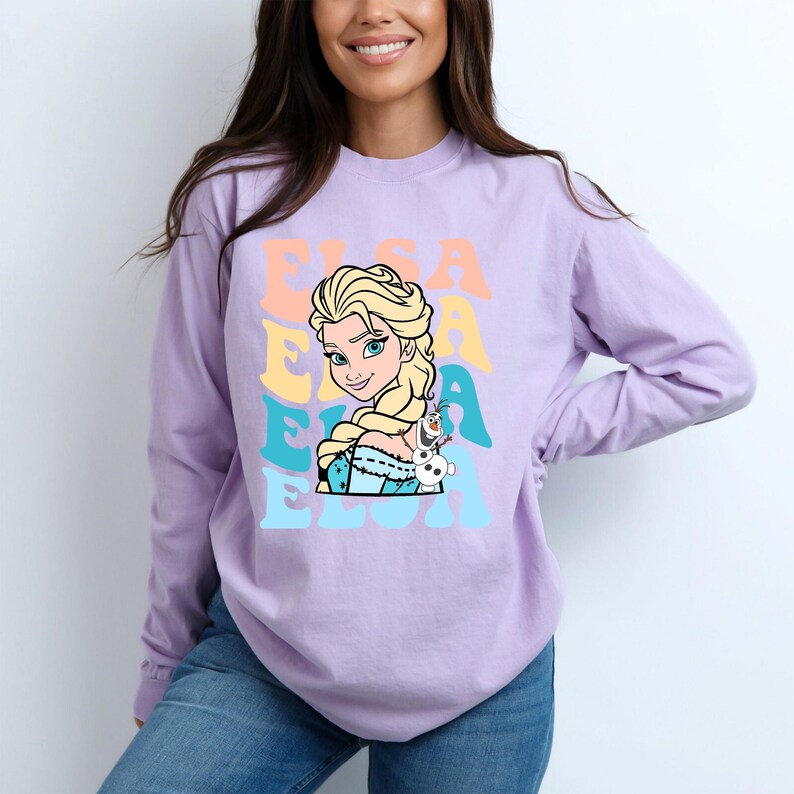 Puede incluir: Una camiseta de manga larga de color lila claro con un gr&aacute;fico de Elsa de Frozen, y el nombre "ELSA" en una fuente retro. Elsa se muestra con cabello rubio y un vestido azul, con Olaf el mu&ntilde;eco de nieve a su lado.