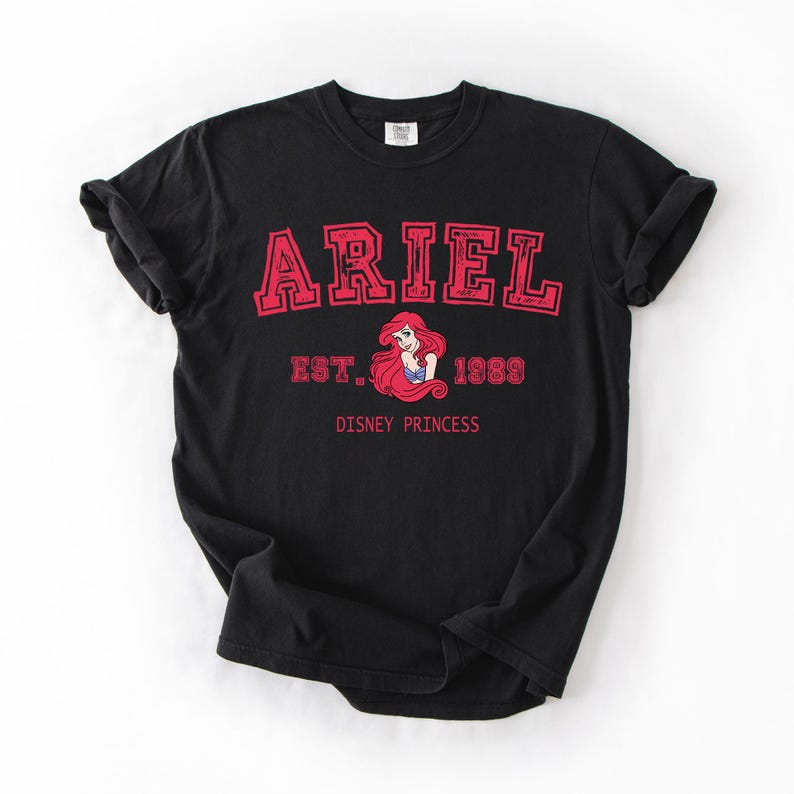K&ouml;nnte beinhalten: Schwarzes T-Shirt mit dem Namen "ARIEL" in roter Schrift im College-Stil. Es enth&auml;lt "EST. 1989" und eine Grafik von Arielle aus Disneys "Arielle, die Meerjungfrau". Darunter steht "DISNEY PRINCESS".