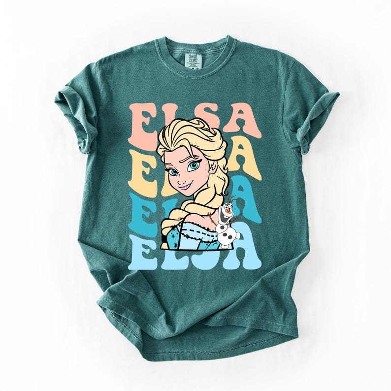 Puede incluir: Camiseta de color verde azulado con un gr&aacute;fico de Elsa de Frozen. El dise&ntilde;o incluye el nombre "Elsa" en una fuente retro con colores pastel. Elsa se representa con cabello rubio, ojos azules y un peque&ntilde;o amigo mu&ntilde;eco de nieve.