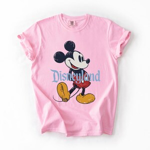 Puede incluir: Camiseta rosa claro con un dise&ntilde;o de Mickey Mouse. El dise&ntilde;o incluye la palabra "Disneyland" en una fuente azul claro. La camiseta tiene mangas enrolladas y cuello redondo.