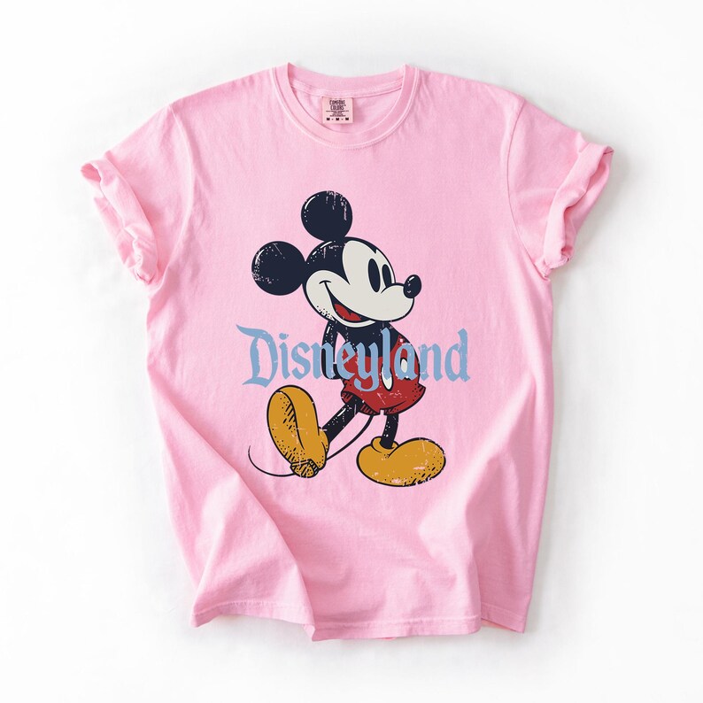 Puede incluir: Camiseta rosa claro con un dise&ntilde;o de Mickey Mouse. El dise&ntilde;o incluye la palabra "Disneyland" en una fuente azul claro. La camiseta tiene mangas enrolladas y cuello redondo.