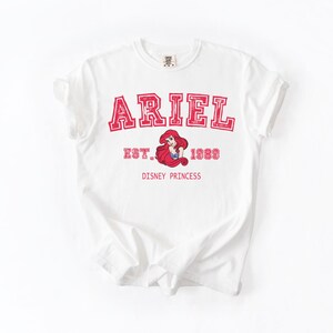 K&ouml;nnte beinhalten: Wei&szlig;es T-Shirt mit dem Namen "ARIEL" in roten Blockbuchstaben mit roter Umrandung. Darunter befindet sich eine Grafik von Ariel, der Disney-Prinzessin, mit dem Text "EST. 1989 DISNEY PRINCESS".