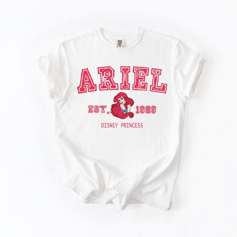 K&ouml;nnte beinhalten: Wei&szlig;es T-Shirt mit dem Namen "ARIEL" in roten Blockbuchstaben mit roter Umrandung. Darunter befindet sich eine Grafik von Ariel, der Disney-Prinzessin, mit dem Text "EST. 1989 DISNEY PRINCESS".