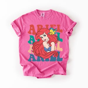 Op de afbeelding: Een roze t-shirt met een afbeelding van Ariel uit De Kleine Zeemeermin. Het woord "ARIEL" staat in kleurrijke retro letters. Ariel heeft rood haar en een blauwe schelp top, met een gele vis op haar hoofd.