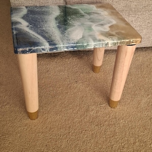 May include: A small, square side table with a resin top depicting an ocean scene in shades of blue, green, and white. The table has light-coloured wooden legs with gold-coloured metal tips. The table is approximately 46 cm long.
