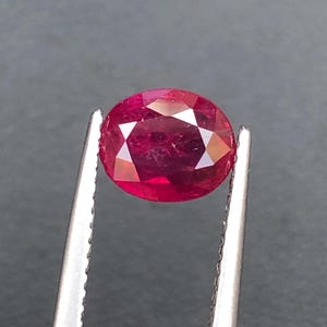 Puede incluir: Una gema de corte ovalado de color rojo intenso con una superficie multifacetada, sostenida firmemente por unas pinzas de metal plateado. La piedra muestra una claridad brillante y un acabado pulido que refleja la luz en sus muchas facetas.