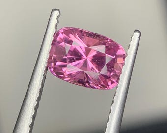 May include: A vibrant pink gemstone with a cushion cut, featuring a faceted surface that reflects light. The stone is held by a pair of silver-toned metal tweezers, showcasing its clear, saturated color and symmetrical geometric pattern.