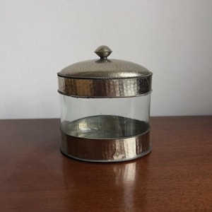 May include: A clear glass jar with a hammered metal lid and base. The lid has a small knob on top. The metal has a silver finish. The jar sits on a wooden surface.