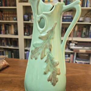 May include: A light green ceramic pitcher featuring a tall, slender body with an irregular, wavy rim and a single handle. The exterior is decorated with two embossed oak leaf motifs in a muted brown tone, positioned vertically along the front.