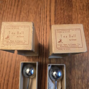 Könnte beinhalten: Zwei Vintage-Tee-Eier von Chase Brass & Copper Co. in originalen beigen Pappschachteln. Jede Schachtel ist beschriftet mit Pol. Chromium Finish & White, No. 90118, Tea Ball by Chase, Waterbury, Connecticut, Made in U.S.A. Die Tee-Eier haben Metallkugeln.