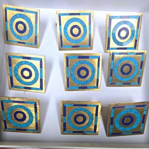 Op de afbeelding: Negen vierkante decoratieve kastknoppen met een gouden afwerking. Elke knop heeft een centrale blauwe cirkel, omgeven door een turquoise cirkel en een blauwe vierkante rand. De knoppen zijn gerangschikt in een witte doos.