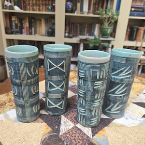 May include: Four tall, cylindrical ceramic tumblers featuring a dark blue-grey glaze with carved, light-colored geometric patterns. Each vessel displays a unique texture, including vertical lines, horizontal bands, X-shapes, and leaf-like motifs.