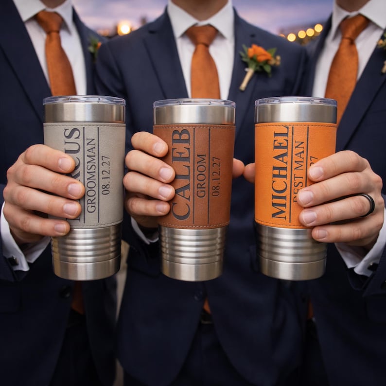 May include: Three stainless steel tumblers with personalized leather sleeves. Each tumbler has a different sleeve color: gray, brown, and orange. The sleeves are engraved with names, titles, and dates, suggesting a wedding or special event.