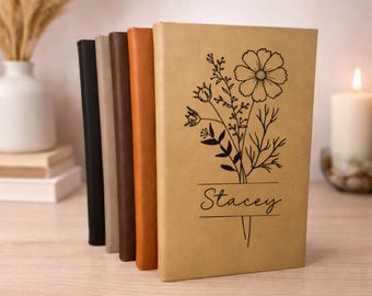 Könnte beinhalten: Eine Reihe von Notizbüchern mit verschiedenfarbigen Einbänden, darunter Schwarz, Beige, Braun und Orange. Das vordere Notizbuch ist beige mit einem Blumendesign und dem Namen "Stacey". Eine brennende Kerze und eine Vase sind im Hintergrund.