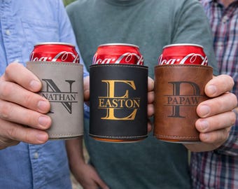 Könnte beinhalten: Drei personalisierte Getränkehalter aus grauem, schwarzem und braunem Kunstleder, die jeweils eine rote Limonadendose halten. Die Halter zeigen individuelle Initialen und Namen: "NATHAN", "EASTON" und "PAPAW", ideal als Geschenk.