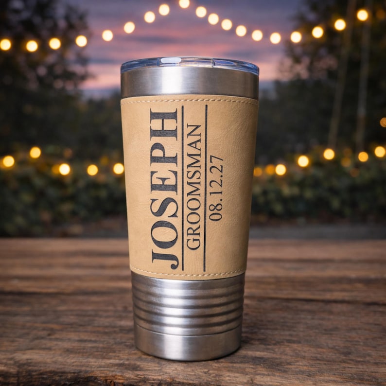 May include: A stainless steel insulated tumbler with a light brown leather wrap. The wrap is engraved with the name "JOSEPH", the title "GROOMSMAN", and the date "08.12.27". The tumbler has a silver lid and a ridged base.