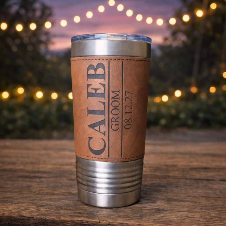 May include: Stainless steel tumbler with a brown leather wrap. The wrap is engraved with the name "CALEB", the word "GROOM", and the date "08.12.27". The tumbler has a clear lid and is sitting on a wooden surface.