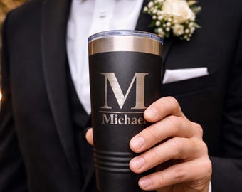 Könnte beinhalten: Ein schwarzer isolierter Becher mit silbernem Rand und Deckel. Der Becher hat einen großen silbernen Buchstaben "M" und den Namen "Michael" darunter eingraviert. Der Becher wird von einer Person in einem schwarzen Anzug und einer Fliege gehalten.