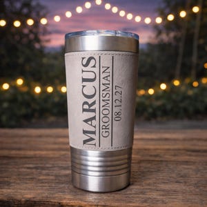 May include: A stainless steel tumbler with a light gray leather wrap. The wrap is printed with the name "Marcus", the title "Groomsman", and the date "08.12.27". The tumbler has a silver lid and base.
