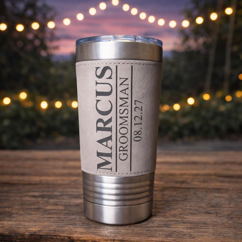 May include: A stainless steel tumbler with a light gray leather wrap. The wrap is printed with the name "Marcus", the title "Groomsman", and the date "08.12.27". The tumbler has a silver lid and base.