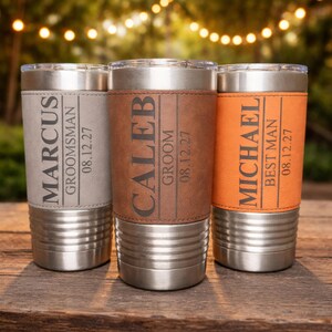 May include: Three stainless steel insulated tumblers with personalized leather sleeves. The tumblers are engraved with names and titles: Marcus, Groomsman; Caleb, Groom; and Michael, Best Man, along with a date.