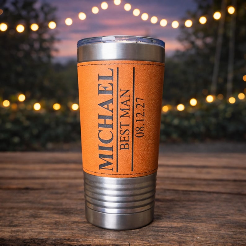 May include: A stainless steel tumbler with an orange leather wrap. The wrap is engraved with the name "MICHAEL", the text "BEST MAN", and the date "08.12.27". The tumbler has a clear lid.