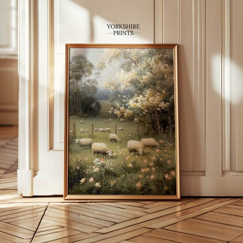 Op de afbeelding: Een ingelijst landschapsschilderij met een pastorale sc&egrave;ne met schapen die grazen in een veld met wilde bloemen. Het kunstwerk heeft een gouden lijst en de tekst "YORKSHIRE PRINTS" is bovenaan zichtbaar. De schildering wordt getoond tegen een witte muur en een houten vloer.