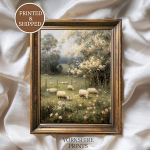 Op de afbeelding: Een ingelijst landschapsschilderij met een pastorale sc&egrave;ne met schapen die in een veld grazen. Het kunstwerk is geplaatst in een goudkleurige houten lijst. Het schilderij toont een groen veld, bomen en een bewolkte lucht. De tekst "PRINTED & SHIPPED" en "YORKSHIRE PRINTS" zijn zichtbaar.