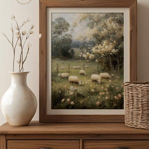 Op de afbeelding: Een ingelijste prent van een pastorale sc&egrave;ne met schapen die in een veld grazen. Het kunstwerk is geplaatst in een houten lijst en toont een weelderige groene weide met bomen en bloemen. De prent wordt getoond op een houten dressoir met een vaas en een mand.