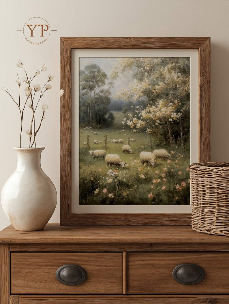 Op de afbeelding: Een ingelijste prent van een pastorale sc&egrave;ne met schapen die in een veld grazen. Het kunstwerk is geplaatst in een houten lijst en toont een weelderige groene weide met bomen en bloemen. De prent wordt getoond op een houten dressoir met een vaas en een mand.
