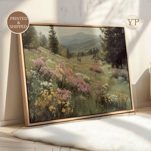 May include: A framed landscape print featuring a vibrant meadow filled with wildflowers in shades of pink, yellow, and white. The artwork depicts a rolling hillside with trees and mountains in the background. The frame is a light wood color.